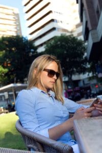 Mujer joven con gafas de sol sentada en una terraza de bar mientras consulta su móvil